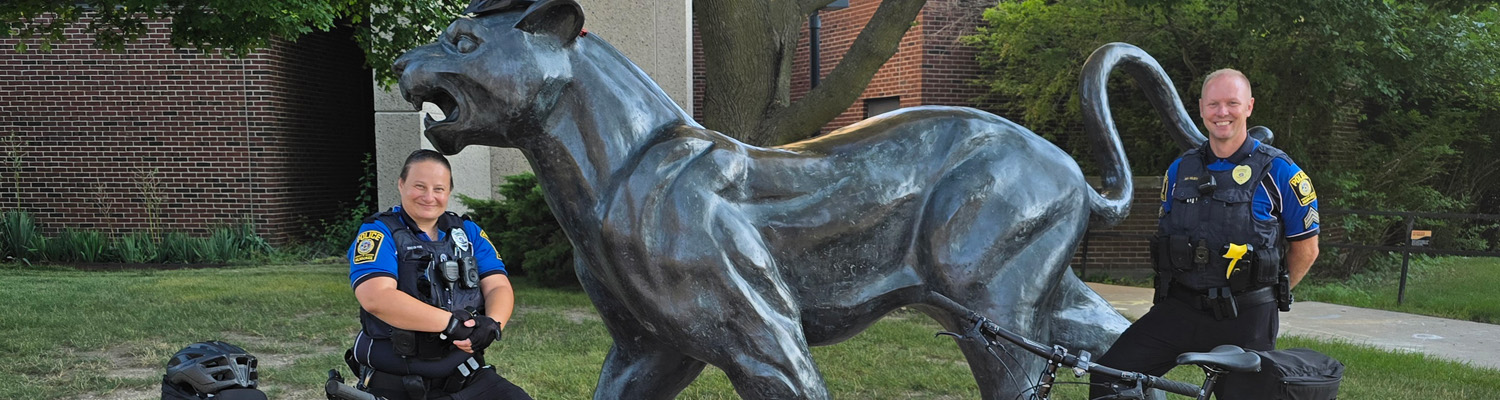 Two police officers posing with bicycles beside a large bronze panther statue on a grassy area with brick buildings in background.