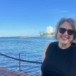 A woman in sunglasses stands on a dock in front of an expanse of blue water
