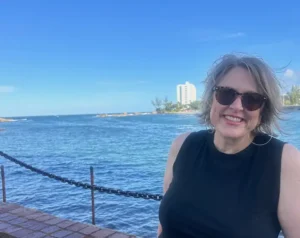 A woman in sunglasses stands on a dock in front of an expanse of blue water