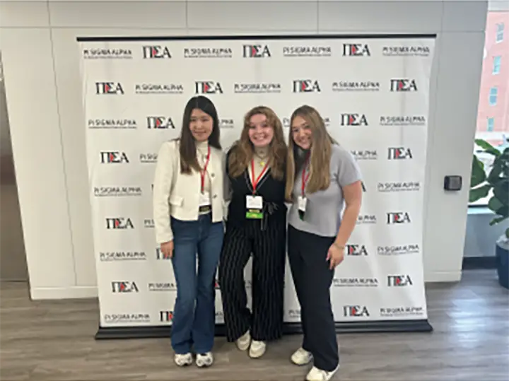 Three young women stand side-by-side in front of a backdrop that reads NCA in repeating letters.