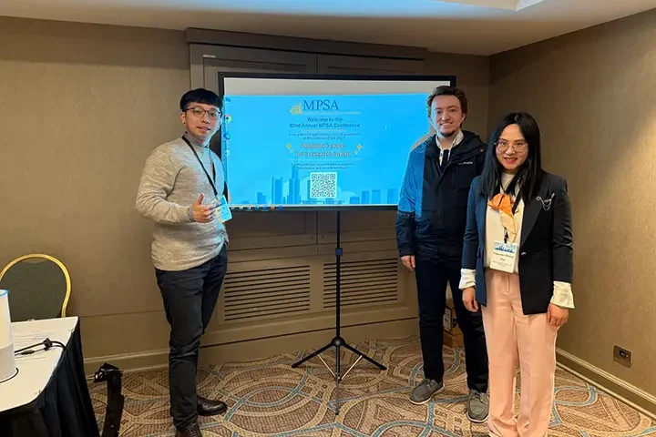 A young man and two friends stand on either side of a projector screen showing an image of the man's research project.