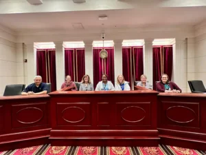 A group of seven people, six women and one man, sit behind a judge's desk in a courtroom with red curtains and carpet.