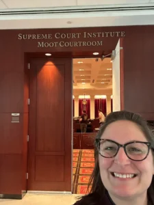 A woman with glasses takes a selfie in front of wood-paneled doors marked with a sign reading Supreme Court Institute Moot Courtroom.