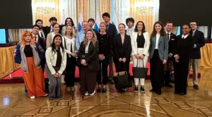 A large group of students stands under brightly-lit chandeliers at the UN Building.