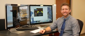 Man smiling at his desk with dual monitors displaying colorful brain scan images