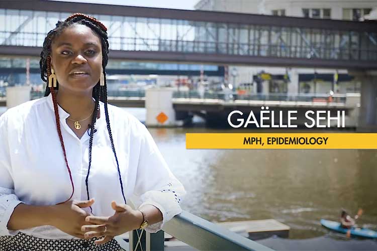 Gaelle Sehi, a student at the Zilber College of Public Health. African-american woman with braids wearing a white blouse. She is standing at a railing of the Milwaukee Riverwalk in downtown Milwaukee. There is a kayaker on the river and Wisconsin Avenue bridge behind her.