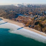 An arial photo of the icy, snow-covered shoreline of Lake Michigan new Atwater Beach, during winter, with downtown Milwaukee rising in the background.