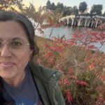 A casual photo of Rachel Goochey with fall leaves on the trees and a waterway behind her. She has long brown hair, glasses, and is smiling.