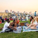 Groups of young men and women sitting on a blanket in a park, with the Milwaukee skyline in the background.