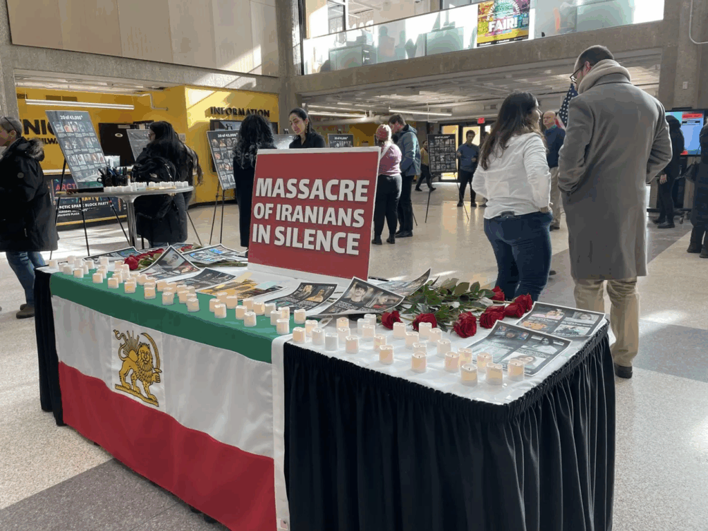 A photo of a table in the UW-Milwaukee Student Union. The table is draped with an Iranian flag. There are candles and red roses on the table and a sign that says "Massacre of Iranians in Silence."