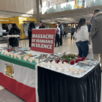 A photo of a table in the UW-Milwaukee Student Union. The table is draped with an Iranian flag. There are candles and red roses on the table and a sign that says "Massacre of Iranians in Silence."