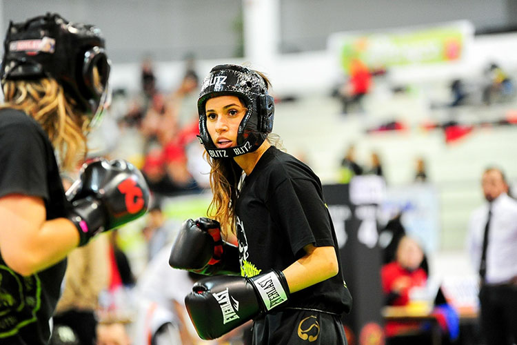 Two women boxing, with a coach and audience watching in the background.