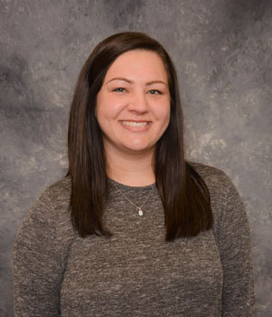 A photo of a smiling woman with long brown hair, wearing a brown sweater.