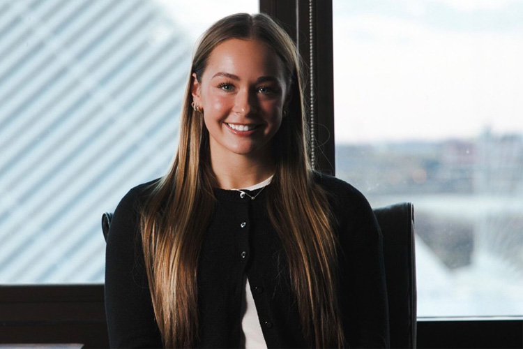 Angelica Refinski, a smiling woman with long dark blonde hair, standing in front of a window.