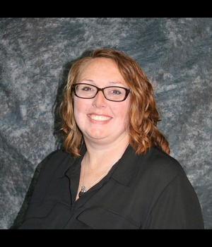 Headshot of Brenda Johansen smiling, wearing glasses and a black blouse, with shorty wavy auburn hair against a textured gray background.