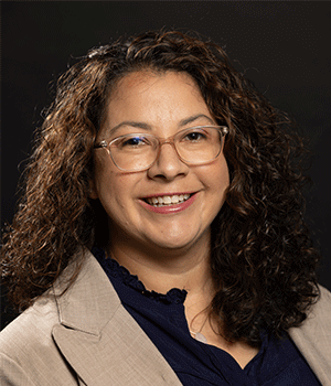 Headshot of Dr. Brooke Slavens smling, wearing glasses and a beige blazer over a navy blue blouse, with curly dark hair against