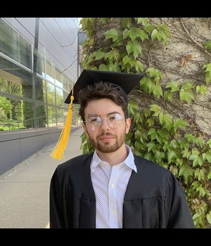 Headshot of Dakota DeLaruelle wearing glasses and a graduation cap and gown, standing outdoors near a building and ivy-covered wall.