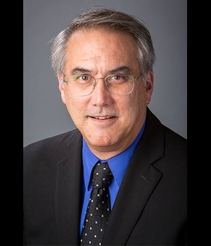 Headshot of Dr. Roger Smith wearing glasses, a dark suit, blue shirt, and patterned tie, against a gray background.