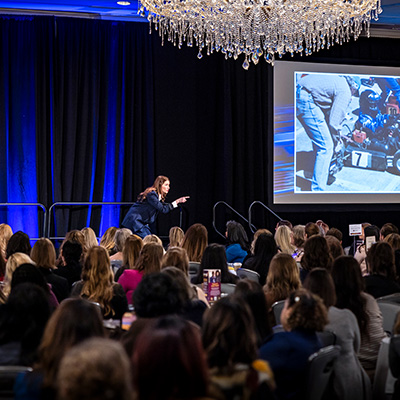 Julia Landauer, a keynote speaker, gestures energetically on stage in front of a large audience, with a presentation slide showing a racing scene projected behind her.