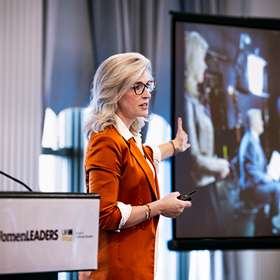 Katrina Cravy, a woman in an orange blazer, presents beside a screen, gesturing toward an image as she speaks at a conference podium labeled “Women Leaders.”