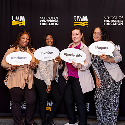 Four women stand together in front of a black "UWM School of Continuing Education" backdrop. They are smiling and holding white speech-bubble signs that read "#Recharge," "#Passion," "#Leadership," and "#Purpose." Each sign also features the "WomenLEADERS" logo at the bottom.