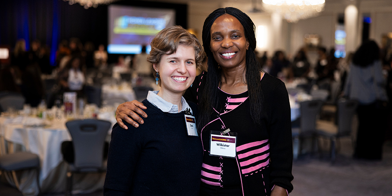 Two conference attendees posing together in a banquet hall with round tables and event participants visible in the background.
