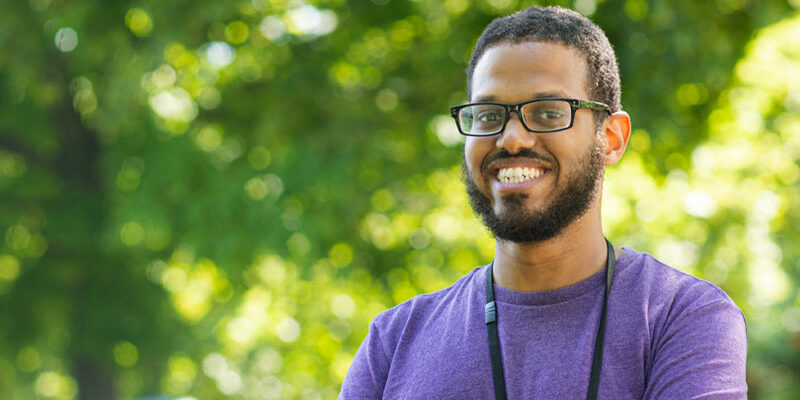 Portrait of Nadir Carlson (black man), a Master's of Social Work student in a purple tee-shirt with lush, green trees in the background.