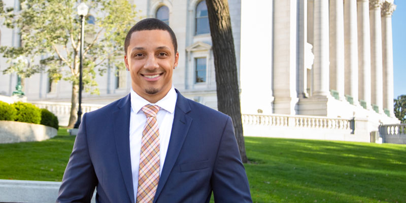 A young student (black man) wearing a suit and tie, standing in front of the Wisconsin State Capitol building.