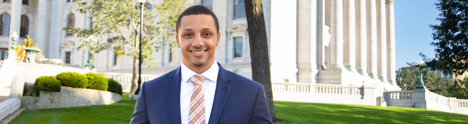A young student (black man) wearing a suit and tie, standing in front of the Wisconsin State Capitol building.