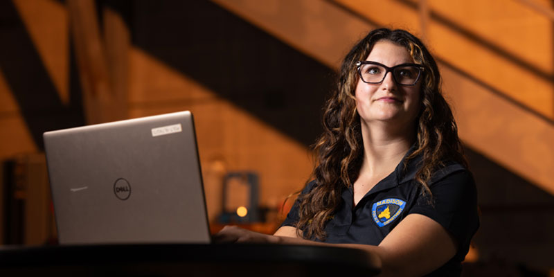 A female crime analyst sits at a table in a glowing, yet dark room looking up and away from her computer screen to the corner of the room.