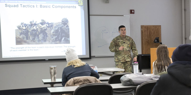 ROTC instructor (white man) giving a presentation to a room of students with a screen in the background that reads: "Squad Tactics I: Basic Components."