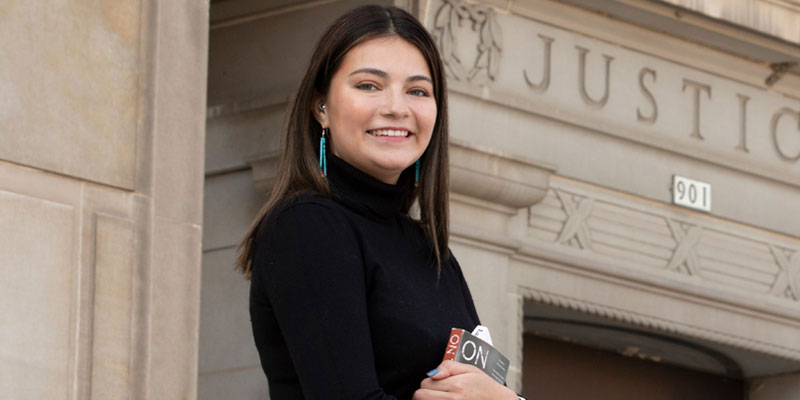 A young woman (Native American) holding a textbook and standing in front of a courthouse building. She is wearing a black turtleneck and dangling turquoise earrings.