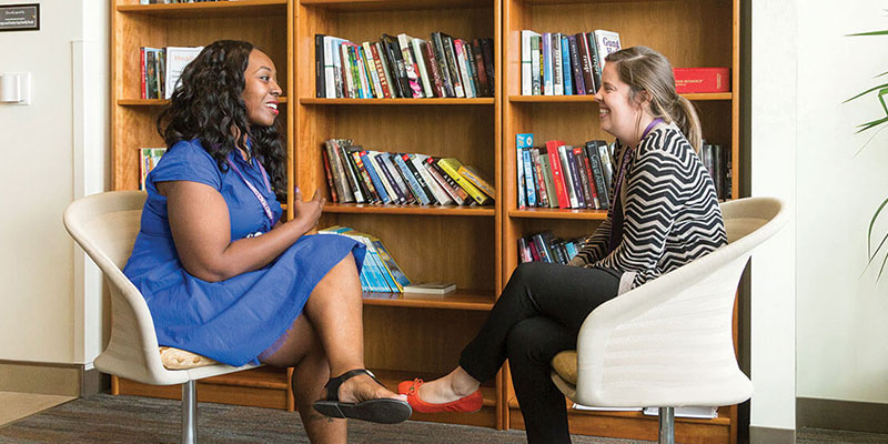 Two female social workers (a black woman and white woman) sitting in modern swivel chairs across from each other in front of three wooden bookshelves. They are smiling and talking.