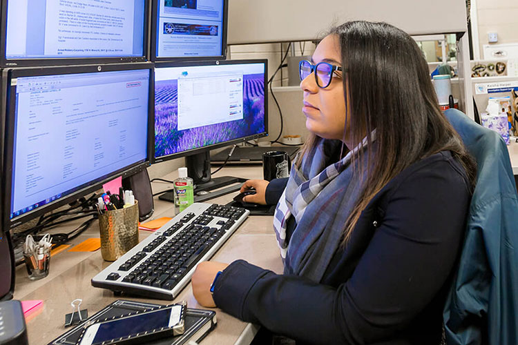 An alumna (multi-racial woman) of the Master's Degree in Criminal Justice & Criminology program who's a crime analyst at the Milwaukee police department. She's sitting in front of four computer screens.