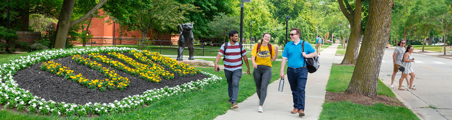 Three students (a man of South Asian descent, a white woman, and a white man) walking together near the Panther Statue and UWM flower garden.