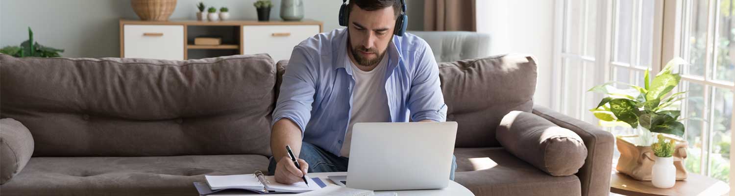 Student (white male) wearing headphones and sitting on a couch with a laptop and writing down an assignment.