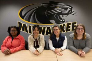 Student Gerontology Association shows four women sitting at a desk in front of wall decal of UW-Milwaukee mascot Pounce Panther.