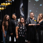 Group of female (white, Asian, Latina) students receiving an award on stage below a gold and black balloon arch.