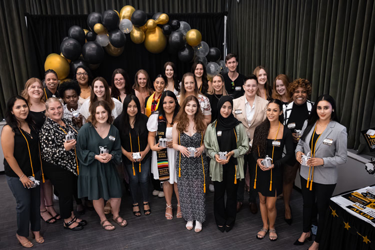 Group of diverse criminal justice & criminology graduating university students posing beneath a black and gold balloon arch.