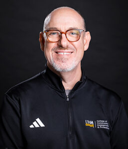 Professional headshot (white male) of individual wearing glasses and a black sporty shirt with logos.