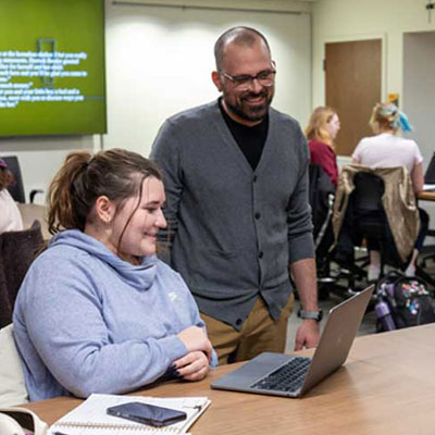 Social work student (white female) sits with a laptop and chats with the instructor (white male) in a classroom setting.