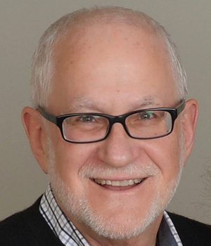 Older adult male wearing glasses and checkered shirt with sweater.