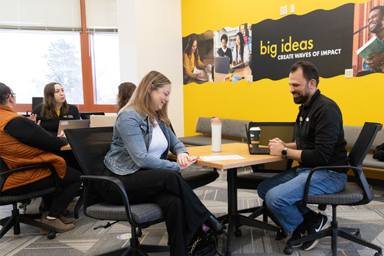 BSW students and academic advisors chatting at tables in front of a brightly colored gold wall.