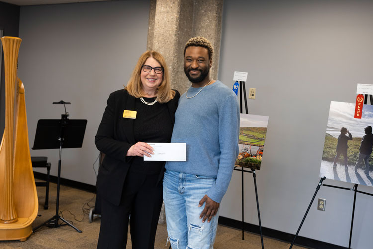 Professor (white female) and student (black male) posing in front of photos on easels. The professor is handing the student an envelope containing an award for his photo.