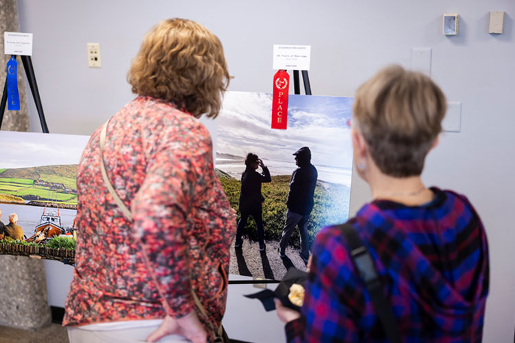 The backs of two women viewing a photo on an easel. The photo shows a woman and a man in a long, dark shadow above a beach while the water shimmers beyond and the sky is brightly lit. A red second place ribbon hangs from the easel and the photo, titled "43 years of marriage."
