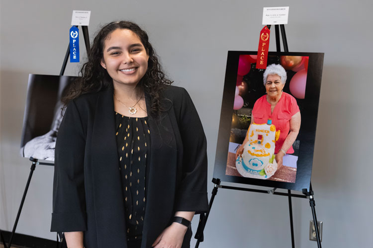 Student dressed up (Latina female) wearing a black blazer, blouse, and a gold sun-shaped necklace while standing in front of a photo on an easel that shows an older adult holding a birthday cake.