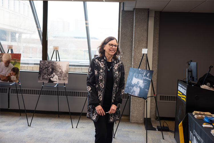 An older adult photographer (white female) smiles and approaches a podium for recognition in a room filled with natural light and photos on easels.
