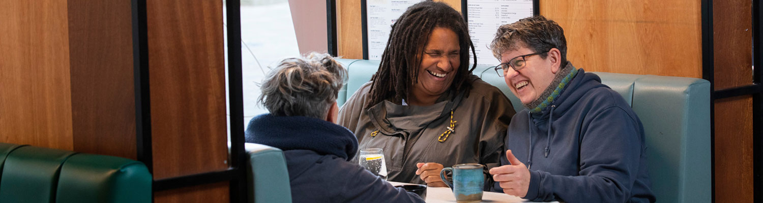 Three older adult friends (one black, two white) sitting in a cafe booth laughing.
