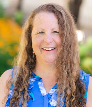Outdoor photo of woman standing in front of an orange flowered tree wearing a blue sleeveless blouse.