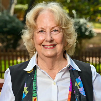 Outdoor headshot of older adult female smiling and wearing a shirt with a colorful scarf draped around her neck.